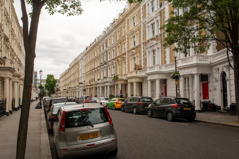 A street view of elegant Victorian-style terraced houses with white and beige facades, detailed cornices, and bay windows. The row of buildings is lined with neatly trimmed hedges and small trees, creating a lush, green border along the pavement. Parked cars of various makes and colors, including grey, black, and bright yellow, fill the street in front of the houses. The sidewalk features traditional black lampposts and is shaded by mature trees with dense foliage, adding a natural canopy over the area. The sky overcast with grey clouds suggests a mild, cloudy day. This scene exemplifies well-maintained urban residential landscaping and street-level garden care, with no visible gardening tools or active maintenance taking place, but it underscores the importance of regular garden and outdoor space upkeep that companies like Gardeners Earls Court provide to maintain such tidy and attractive streetscapes.