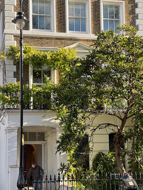 A well-maintained residential garden scene in a city setting featuring a white-painted front porch with decorative columns, lush green climbing plants and trees partially obscuring the view of the building facade, which consists of a brick exterior with large sash windows. In the foreground, a black vintage-style street lamp stands beside a black wrought iron fence that encloses the small front garden. The lawn area is neatly trimmed, with patches of grass visible near the base of the trees and garden beds filled with leafy foliage. A person dressed in dark clothing is seen tending to the garden near the entrance, possibly engaged in gardening or maintenance work. The environment is illuminated by soft daylight, highlighting the textures of the wet soil, trimmed leaves, and the decorative architectural details of the building. This scene exemplifies professional gardening and garden maintenance activities conducted by Gardeners Earls Court, emphasizing landscaping work, hedge trimming, and lawn care in an urban residential setting.