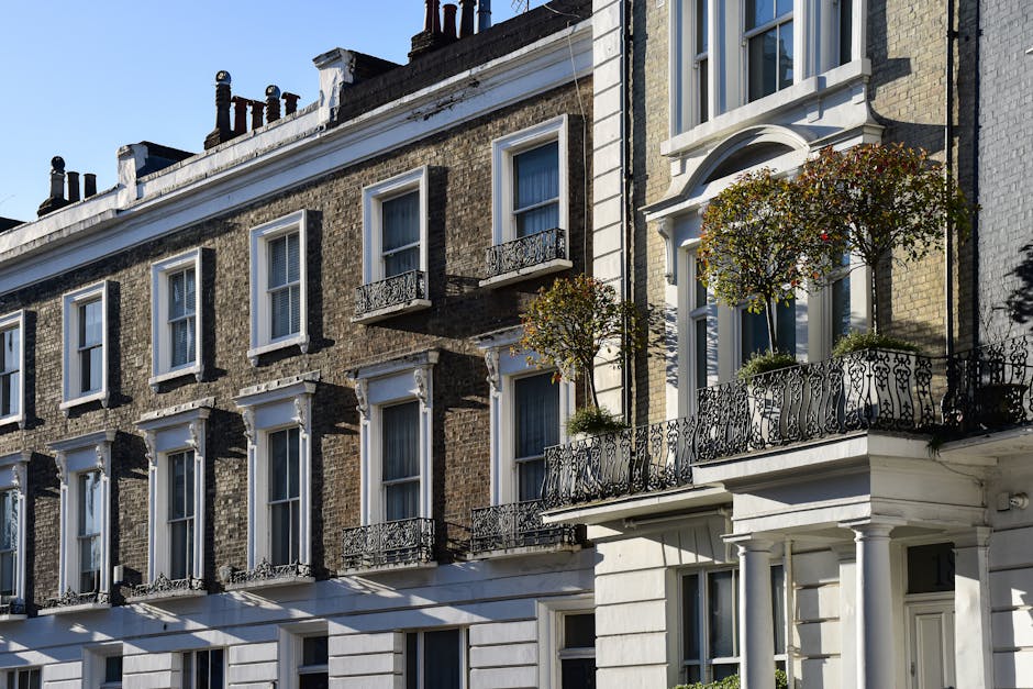 A street view of elegant Victorian-style terraced houses with white and beige facades, detailed cornices, and bay windows. The row of buildings is lined with neatly trimmed hedges and small trees, creating a lush, green border along the pavement. Parked cars of various makes and colors, including grey, black, and bright yellow, fill the street in front of the houses. The sidewalk features traditional black lampposts and is shaded by mature trees with dense foliage, adding a natural canopy over the area. The sky overcast with grey clouds suggests a mild, cloudy day. This scene exemplifies well-maintained urban residential landscaping and street-level garden care, with no visible gardening tools or active maintenance taking place, but it underscores the importance of regular garden and outdoor space upkeep that companies like Gardeners Earls Court provide to maintain such tidy and attractive streetscapes.